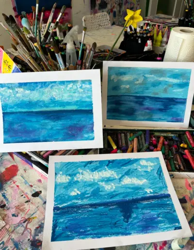 Auf einem Ateliertisch mit vielen Malutensilien stehen drei kleinere Kunstwerke. Die Motive zeigen unterschiedliche Ansichten von Meer und Himmel in blau-weissen Farbtönen.