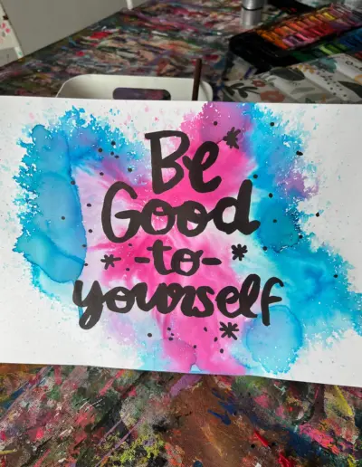 Auf einem weissen Blatt Papier mit blau und rosa Farbklecksen steht in schwarzen Lettern 'Be Good To Yourself'.