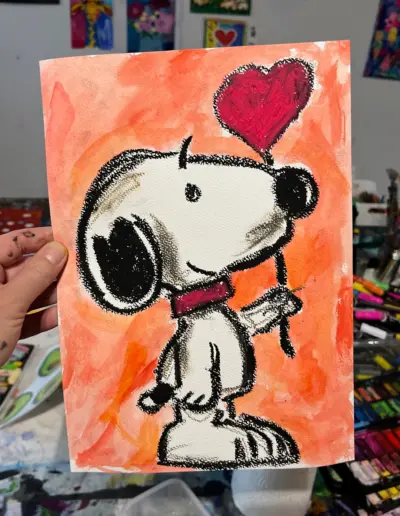 Eine Hand hält ein Blatt Papier, auf der eine Kreidezeichnung von der Figur Snoopy mit einem Herzballon in der Hand gemalt ist.
