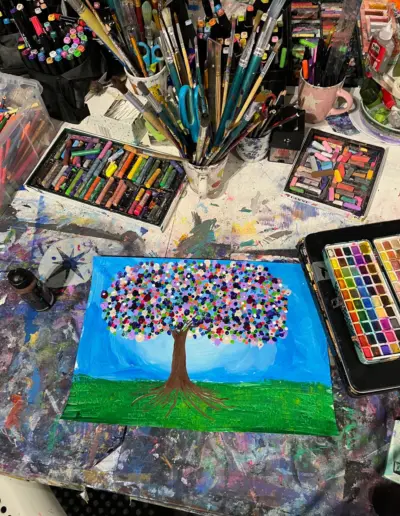 Auf einem Ateliertisch liegt ein Kunstbild, umgeben von vielen Malutensilien wie Kreide, Aquarellkasten, Pinsel, etc. Das Kunstbild zeigt einen farbenfrohen Baum auf einer grünen Wiese vor blauem Himmel.