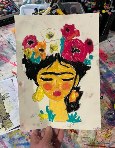 Eine Hand hält ein Zeichenblatt mit einer farbenfrohen Darstellung von Frida Kahlo, gemalt von einer Workshop-Teilnehmerin in Ilkas Malschule.