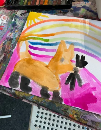 Auf einem Maltisch liegt ein buntes Aquarellbild eines Künstlerkindes, neben dem Bild steht ein Aquarellkasten.