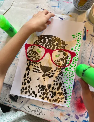 Ein Künstlerkind malt ein Bild von einem Leopard mit roter Brille, umgeben von grünen Farbpunkten.