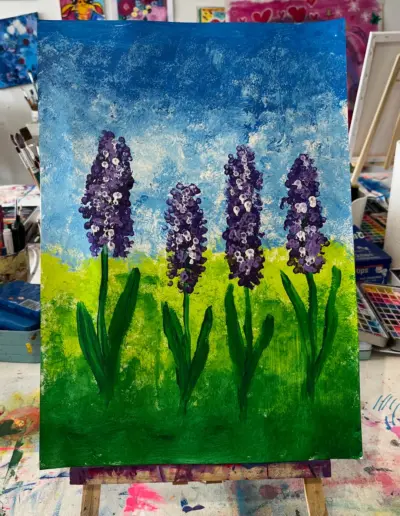 Eine Leinwand mit Acrylmalerei auf einer Tischstaffelei. Das Motiv zeigt vier nebeneinander stehende lila-farbenene Blumen auf einer Sommerwiese mit hell- und dunkelblauem Himmel.