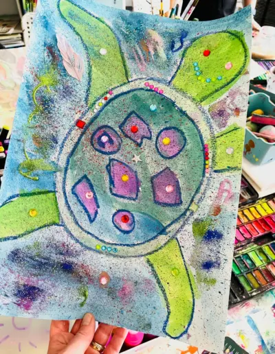 Eine bunte Ölkreide-Zeichnung von einer im Wasser schwimmenden Schildkröte mit grün-violettem Farbmuster auf dem Panzer und bunten Schmucksteinen auf Kopf, Beinen und Panzer.