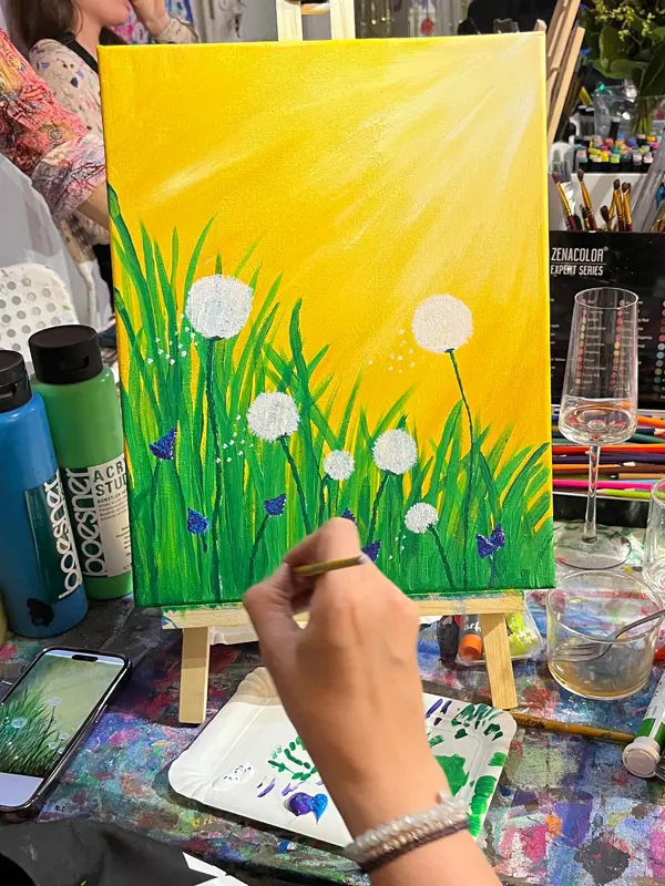 Eine DIN-A4 große Leinwand auf einer kleinen Tisch-Staffelei. Die malerische Arbeit zeigt eine Sommerwiese mit Pusteblumen und Gras vor einem gelben Hintergrund, entstanden im Workshop 'Kunst am Abend und ein Glas Wein'.