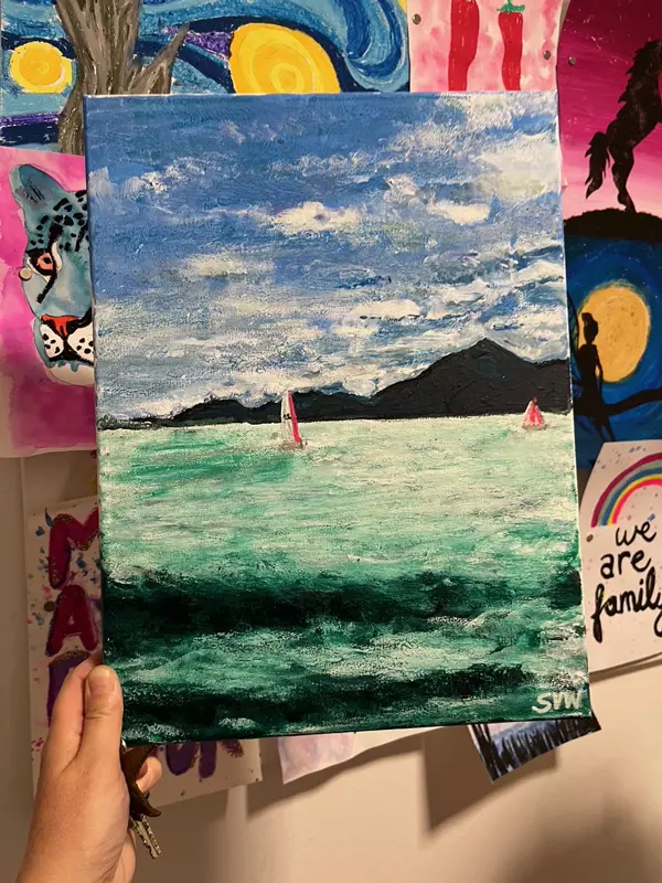 Eine Hand hält eine Leinwand, auf dem ein Meerbild mit Segelbooten auf türkisfarbenem Wasser und dunkleren Küstenbergen im Hintergrund gemalt ist, entstanden im Workshop 'Kunst am Abend und ein Glas Wein' in Ilkas Malschule.