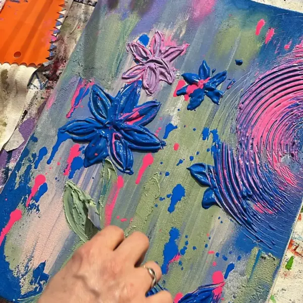 Eine Teilnehmerin von Ilkas Malschule gestaltet mit einer kleinen Kunstspachtel ein Bild mit Blumenmuster und Kreisstrukturen in den Farben blau, violett, grün, rosa und weiteren Farben auf eine Leinwand.