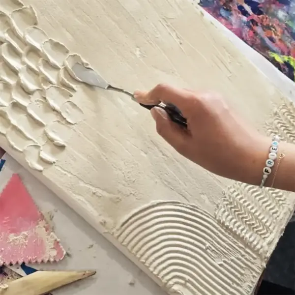 Eine Hand mit einer kleinen Spachtel trägt helle Strukturpaste auf eine Leinwand auf.