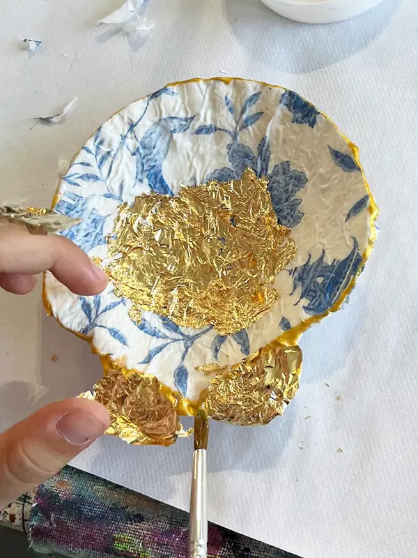 Eine Workshopteilnehmerin gestaltet eine Muschel und trägt vorsichtig etwas Blattgold auf.