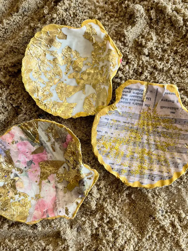 Drei künstlerisch gestaltete Muscheln mit Musiknoten- und Blumenmuster und verziert mit Blattgold, liegen auf einem Sandboden.