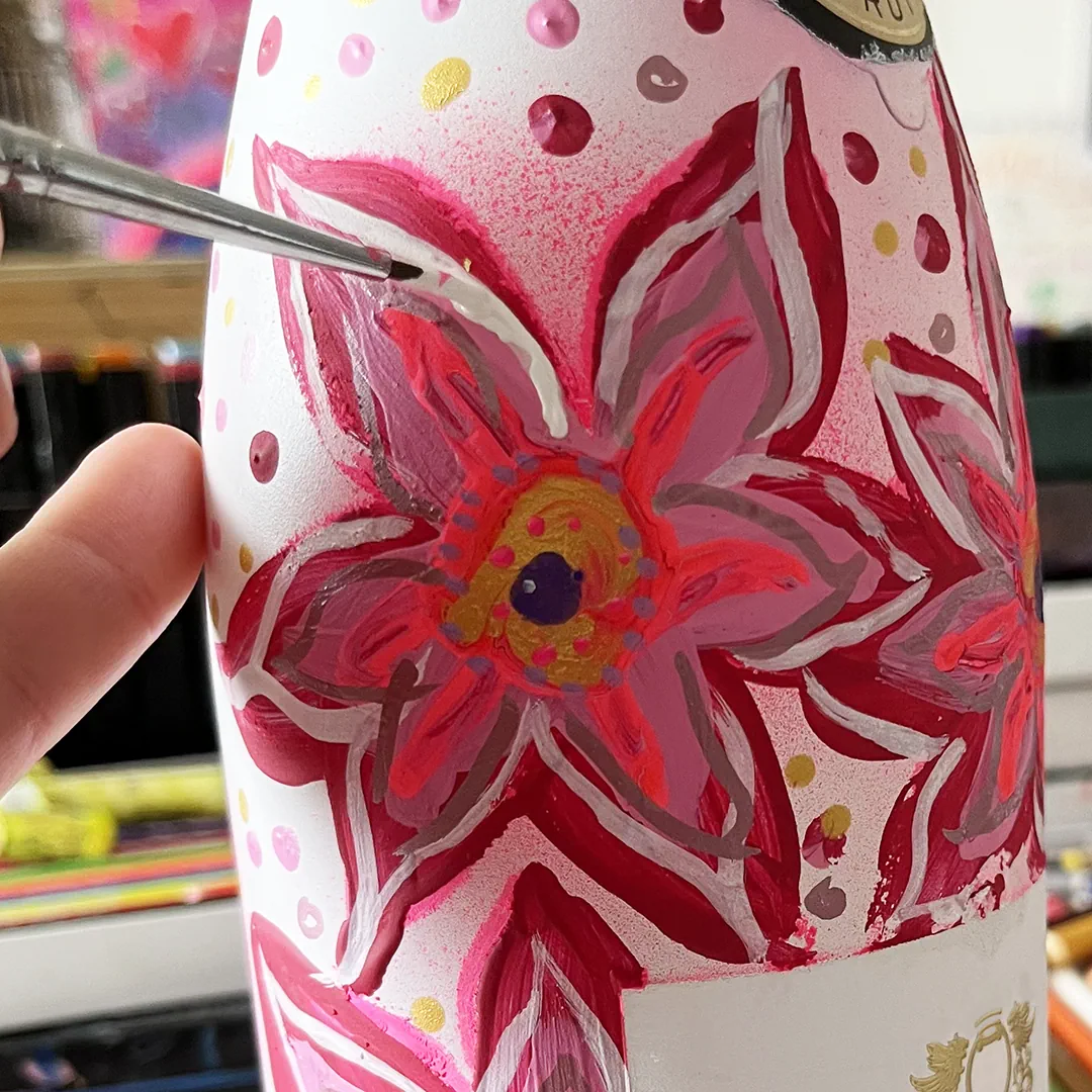 Ein florales Muster in roten, rosa, violetten und goldenen Farbtönen wird mit einem feinen Pinsel auf eine weiß bemalte Champagnerflasche aufgetragen.