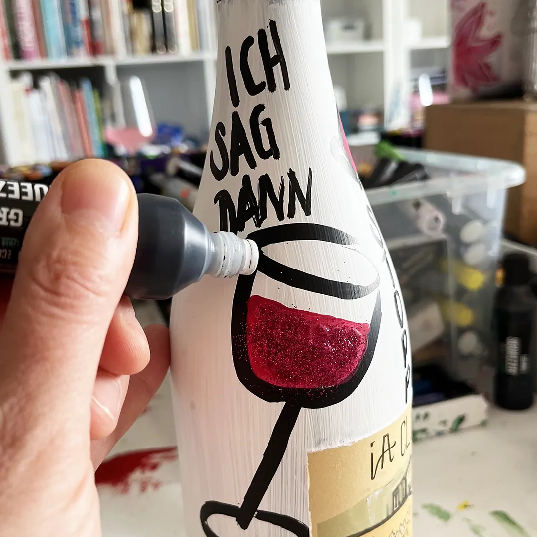 Auf eine weiß angemalte Weinflasche malt eine Workshop-Teilnehmerin mit einem schwarzen Marker einen Spruch und eine Rotweinglas-Illustration.