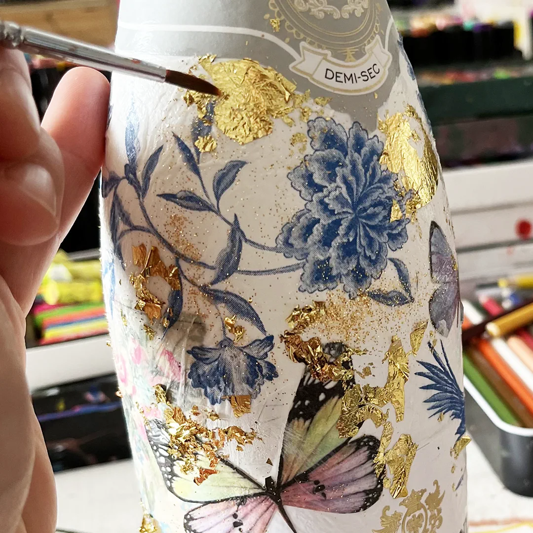 Mit einem feinen Pinsel trägt eine Workshop-Teilnehmerin etwas Blattgold auf eine wunderschön gestaltete Champagnerflasche auf.