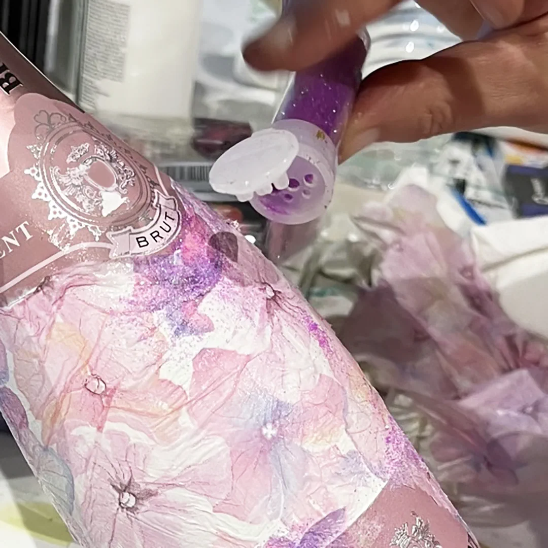 Künstlerische Arbeit aus dem Workshop 'Art on a Bottle' Eine Workshop-Teilnehmerin streut etwas violettes Glitzerpulver auf eine kunstvoll gestaltete Champagnerflasche.