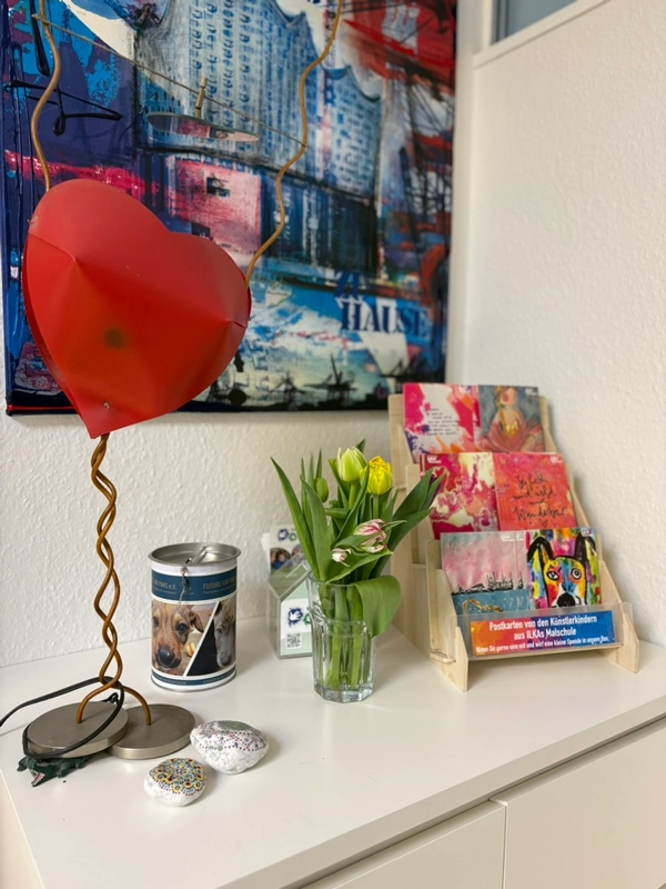 Ein Postkartenständer, zwei Spendenboxen und ein Glas gefüllt mit Blumen stehen auf einer Kommode.